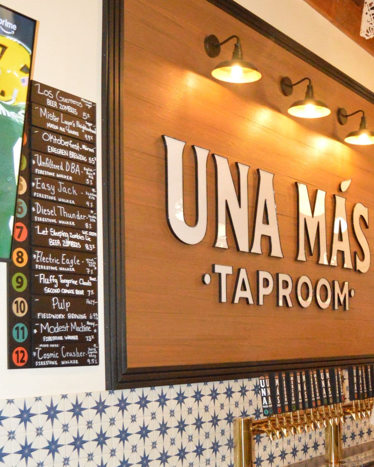 The Una Más Taproom sign over the star tiles, the chalkboard of taps 1 to 12 beside it, brass taps below.
