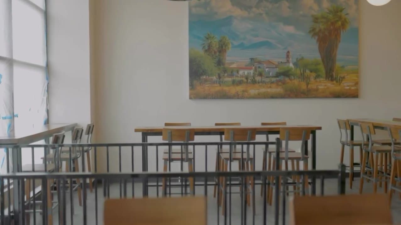 The taproom before opening: long wooden tables and stools under a big painting of a village with palms below the mountains.